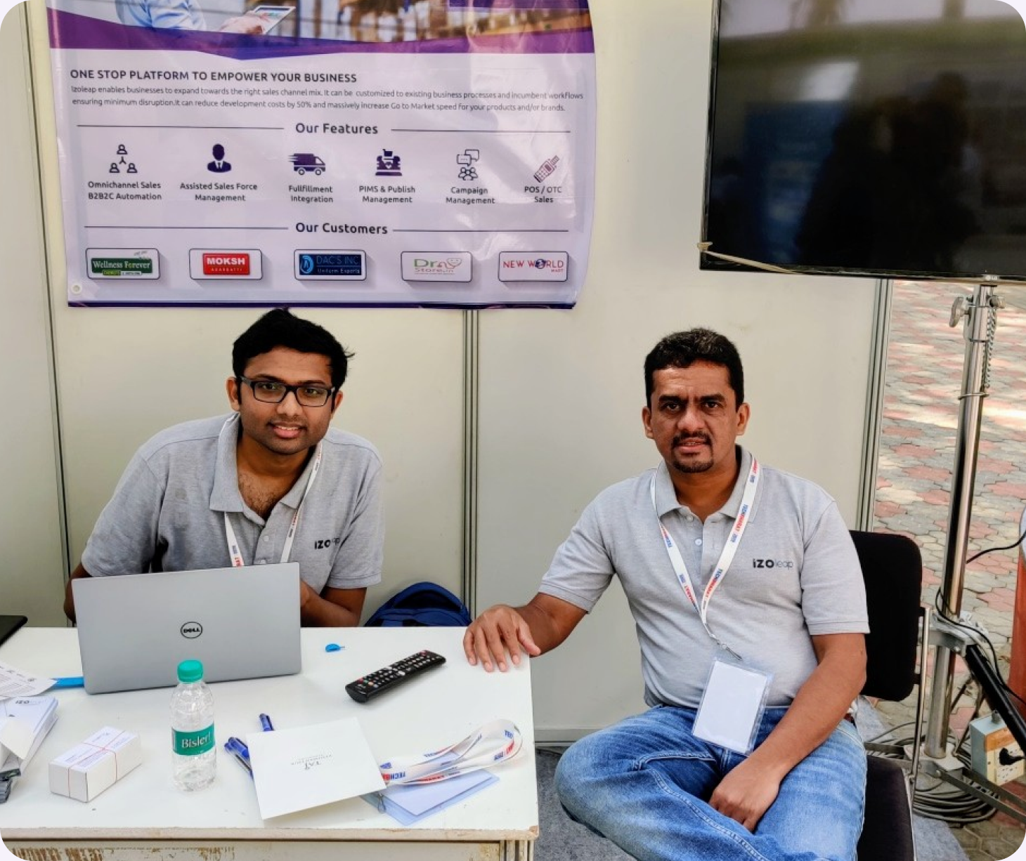 Two Invenzo Labs team members seated at the TechBharat exhibition stand, with their product feature poster and customer logos on the wall behind them.